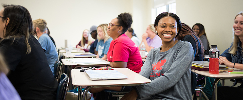 Female student smiling sitting at desk in classroom with other students.